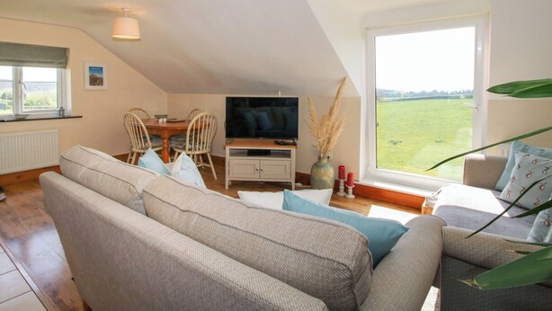 A cozy living room with a sofa set, a dining table with chairs, a TV, and a large window offering a view of a green field. - Mossy Lodge (Shrewsbury)