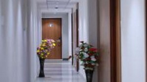 A hotel hallway with a wooden door at the end, a white floor, and two potted plants with flowers. - TM Inn (Kanchipuram)