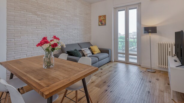 Ein modernes Wohnzimmer mit einem Holzesstisch, weißen Stühlen, einem grauen Sofa mit Kissen, einer Blumenvase und einem Bild an der Wand.