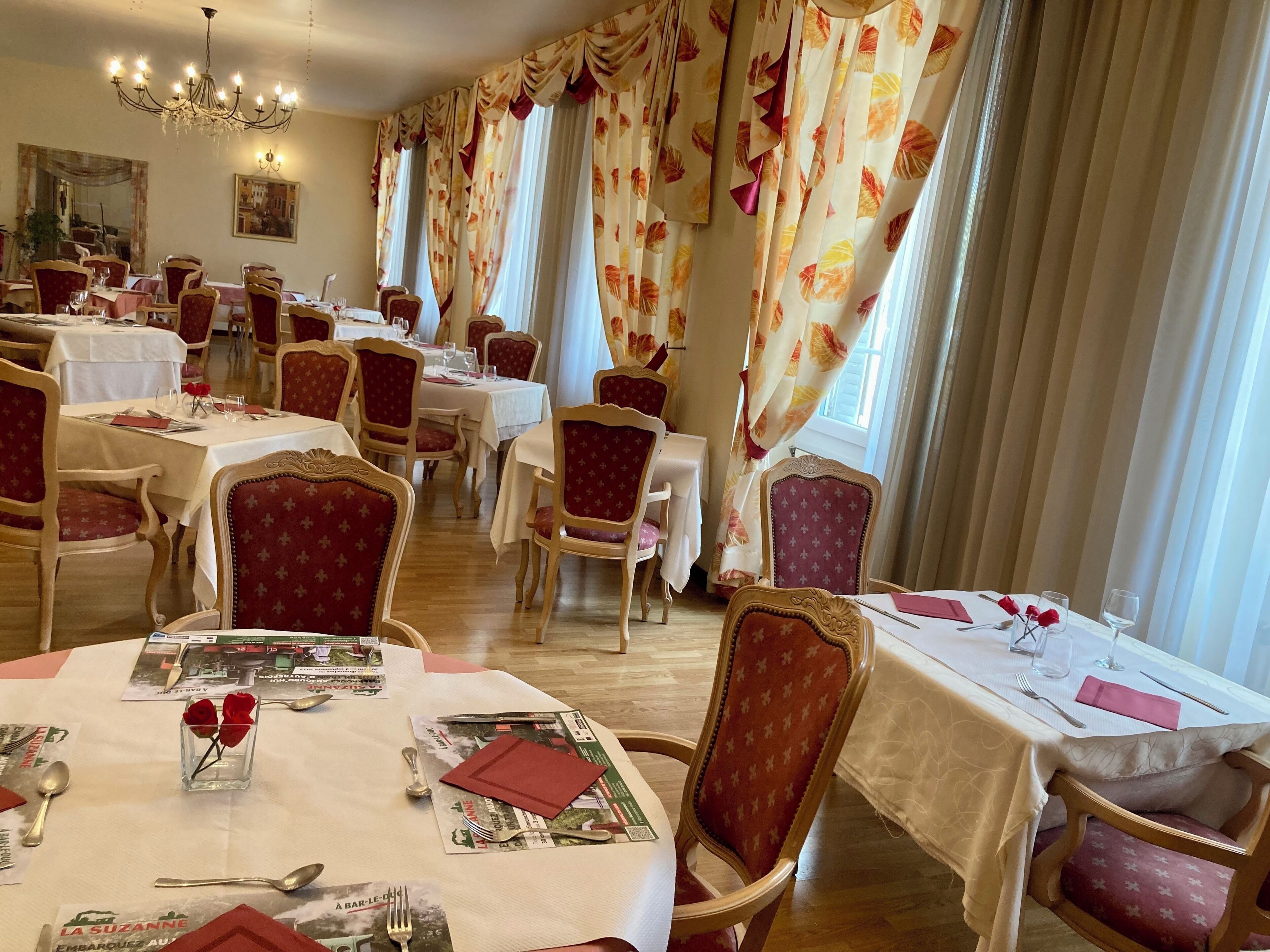 Un comedor con varias mesas dispuestas para comer, cada una con sillas tapizadas en rojo, manteles blancos y cubertería y cristalería ordenadas.