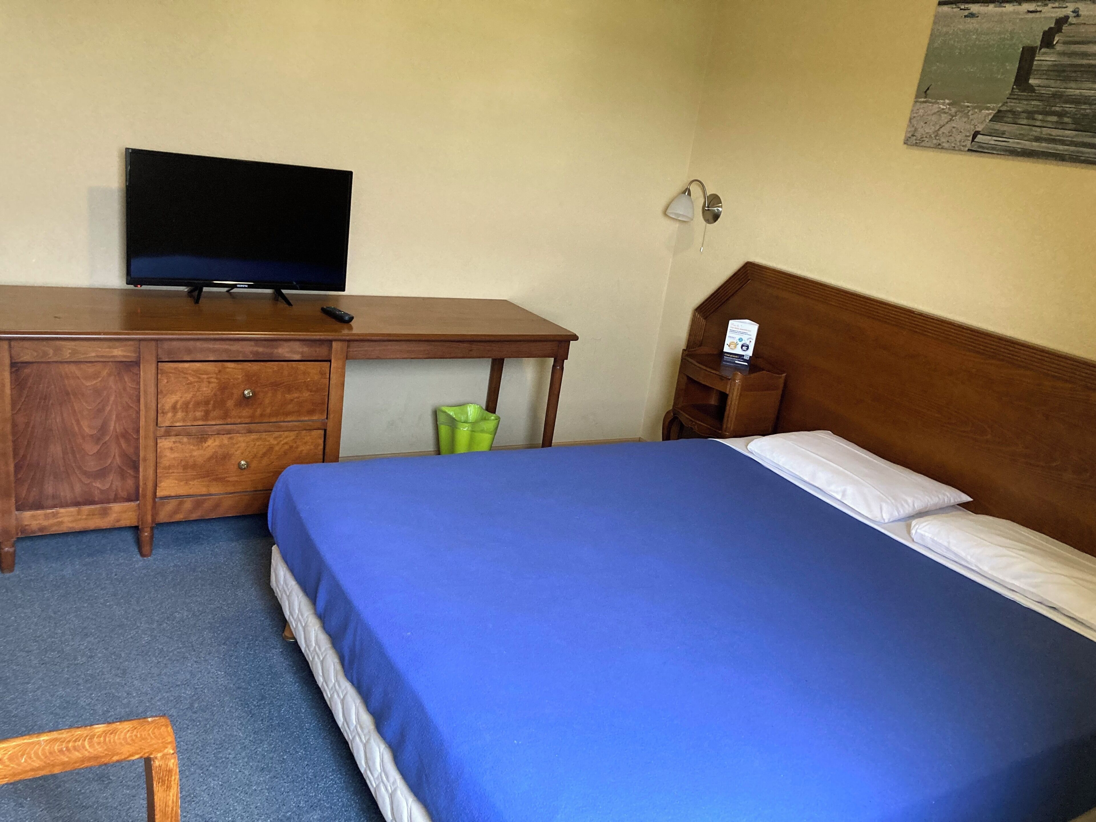Een hotelkamer met een bed, een houten bureau met een televisie, een nachtkastje en een wandlamp.