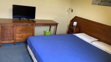 A hotel room with a bed, a wooden desk with a television, a nightstand, and a wall-mounted lamp.