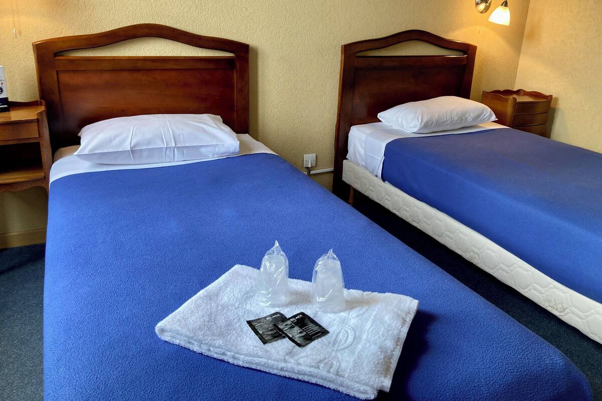 A hotel room with two beds, each with a blue bedspread and white pillows. There is a white towel with a bottle and a packet on it.