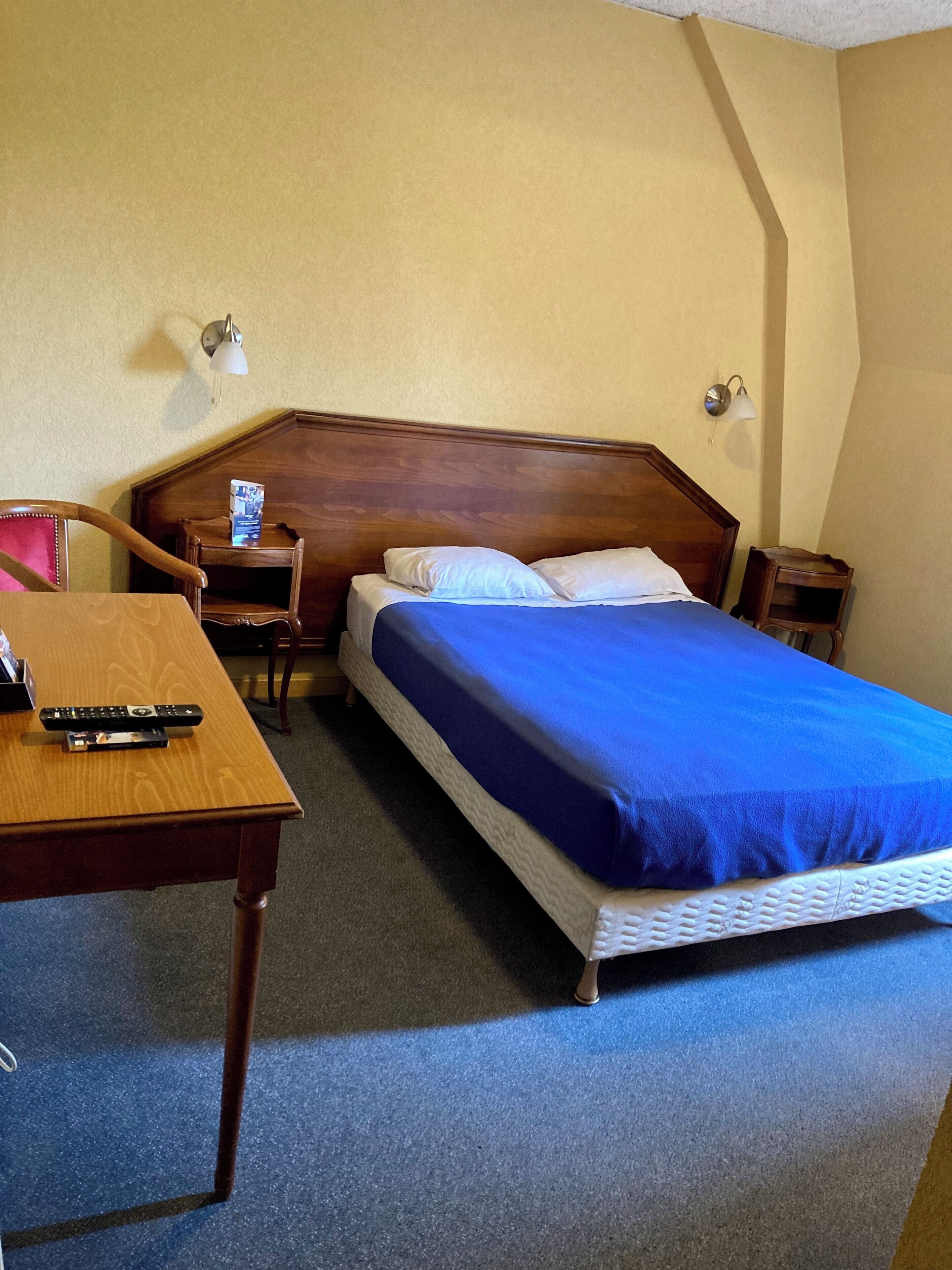 A hotel room with a wooden headboard, a single bed with a blue bedspread, a wooden nightstand, a desk with a remote control, and a chair.