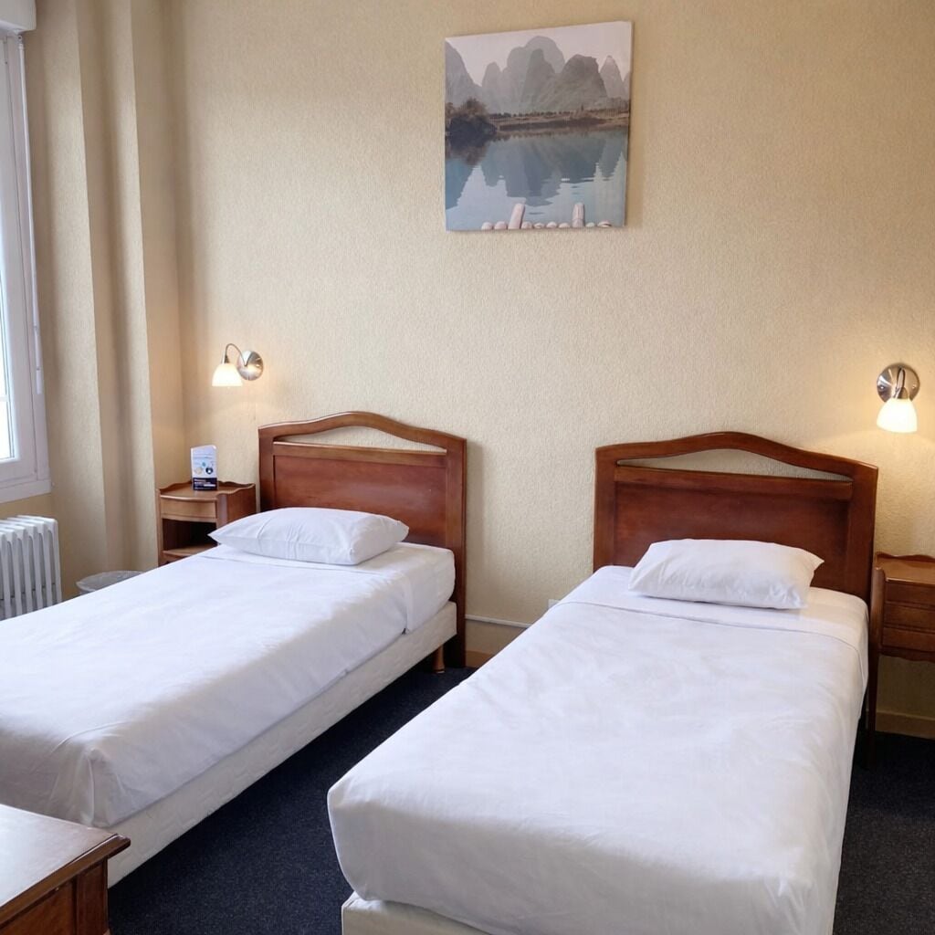 A hotel room with two single beds, a nightstand with a lamp, a radiator, and a painting of a mountain landscape.