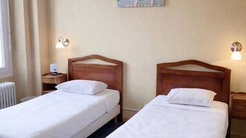 A hotel room with two single beds, a nightstand with a lamp, a radiator, and a painting of a mountain landscape.