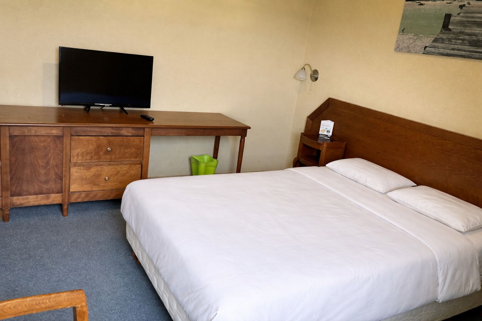 A hotel room with a wooden desk, a television, a bed with white linens, a nightstand, and a wall-mounted lamp.