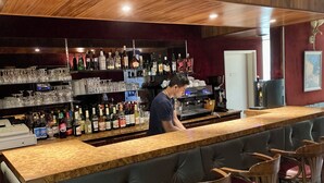 Una zona de bar con una barra alargada, varios taburetes y estantes llenos de botellas y copas.