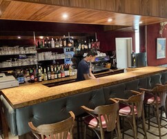 Una zona de bar con una barra alargada, varios taburetes y estantes llenos de botellas y copas.