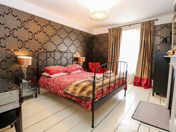 A bedroom with a metal bed frame, a red and beige patterned bedspread, two bedside lamps, a red chair, a small table with a lamp, and a dark-colored wardrobe.