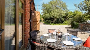 An outdoor dining area with wicker furniture and a glass table, set against a brick building with a wooden door and a view of trees and a gravel path. - The Barnhouse (North Walsham)