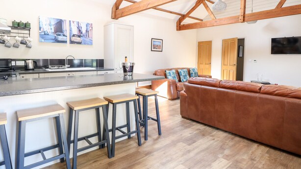 A modern kitchen with a central island, bar stools, a leather sofa, and a TV mounted on the wall. - The Barnhouse (North Walsham)