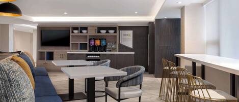 A modern living room with a sofa, a dining table, and bar-style chairs. There is a kitchen area with a TV and storage cabinets.