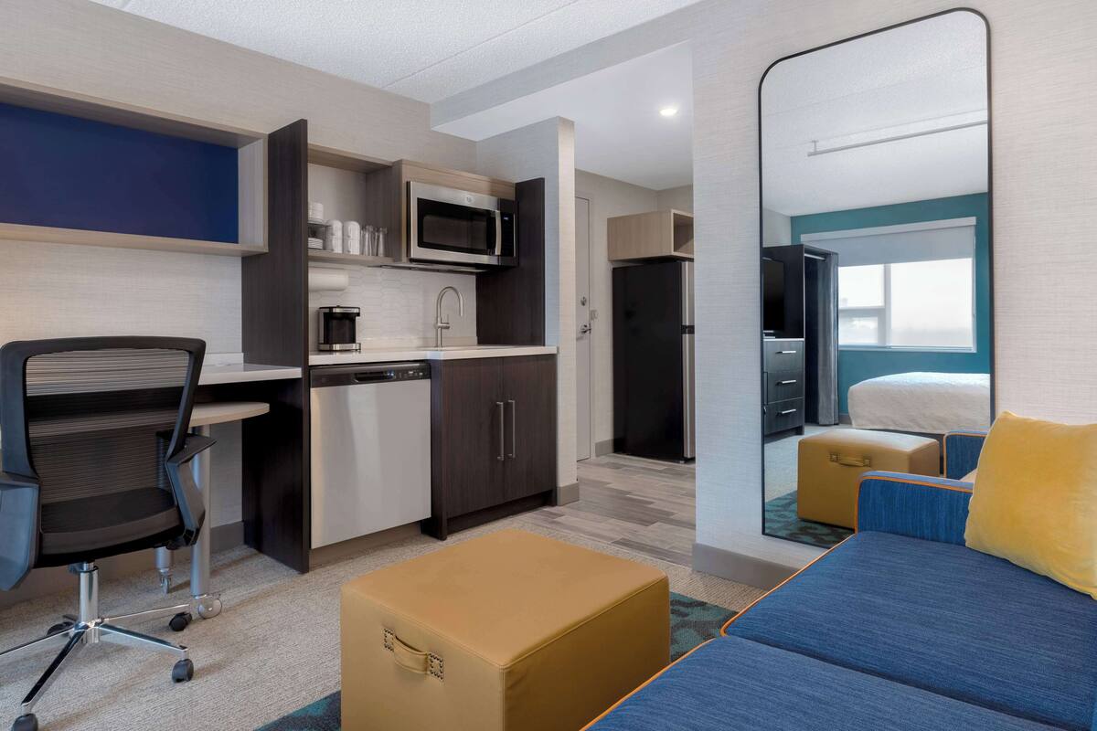 A compact hotel room with a kitchenette, a bed with blue bedding, a large mirror, and a small seating area.