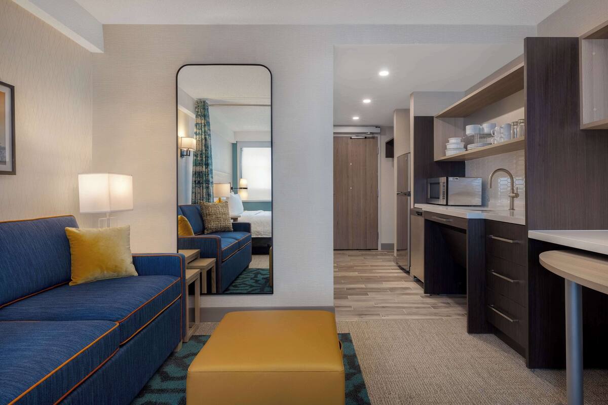 A compact hotel room with a sofa, a small table, a mirror, a kitchenette, and a bathroom visible through an open door.