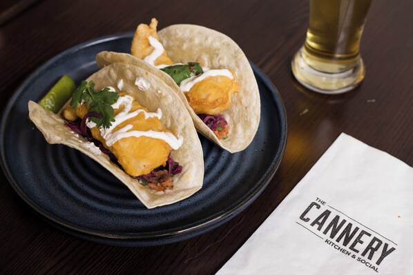 Deux tacos sur une assiette sombre, accompagnés d’une serviette et d’un verre de bière, le tout posé sur une table en bois.