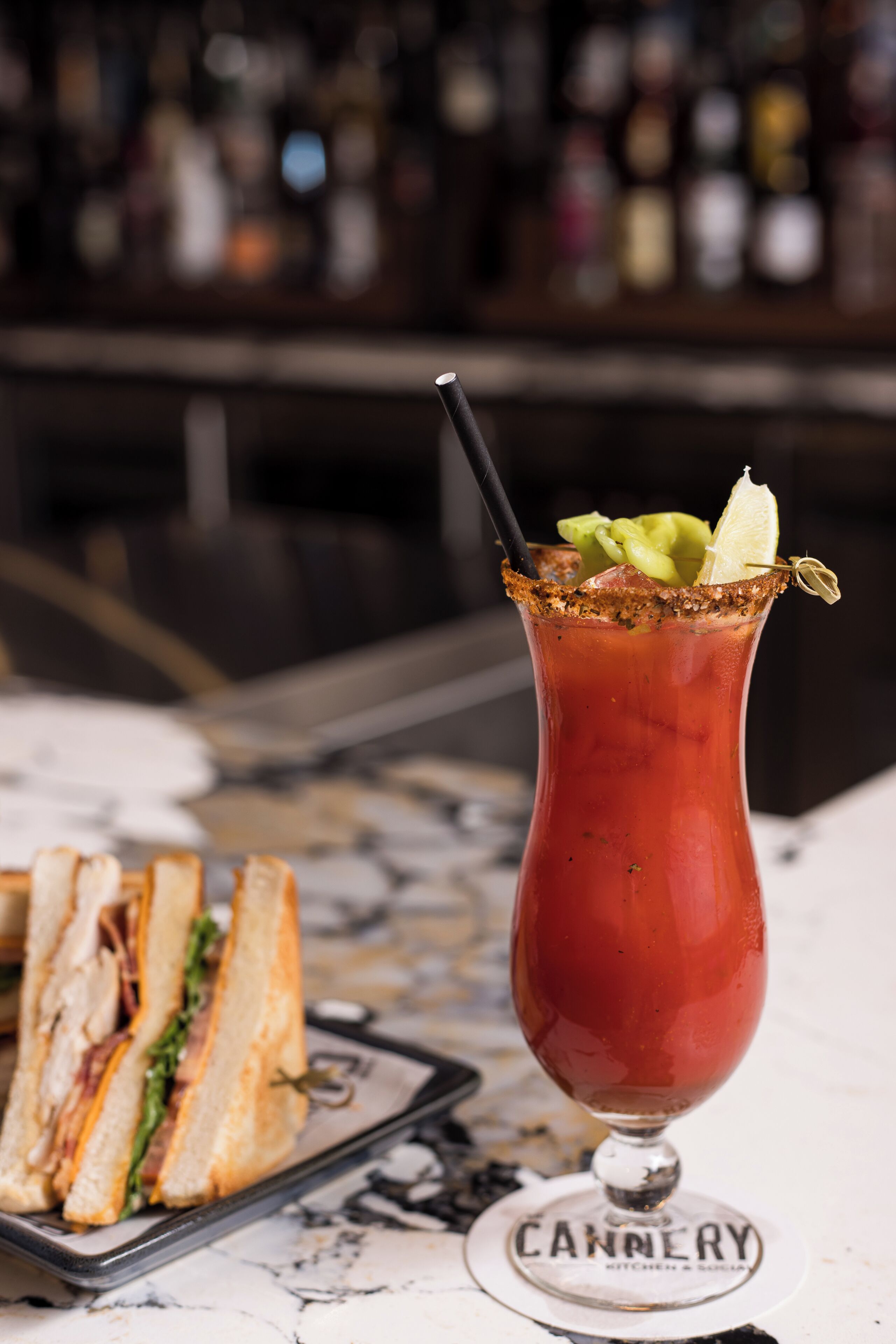 Um cocktail Bloody Mary num copo com borda salgada, decorado com aipo e uma fatia de limão, e um canudo. Um sanduíche está visível num prato ao lado da bebida. O fundo sugere um ambiente de bar com garrafas.