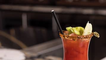 Um cocktail Bloody Mary num copo com borda salgada, decorado com aipo e uma fatia de limão, e um canudo. Um sanduíche está visível num prato ao lado da bebida. O fundo sugere um ambiente de bar com garrafas.