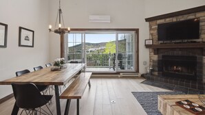 A dining area with a wooden table, black chairs, and a chandelier. There is a fireplace and a television on the wall. - Tremblant Prestige - Manoir 120-6 (Mont-Tremblant)