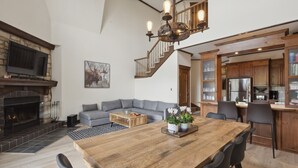 A spacious living room with a fireplace, a dining area with a wooden table, a grey sofa, and a staircase leading to the upper floor. - Tremblant Prestige - Manoir 120-6 (Mont-Tremblant)