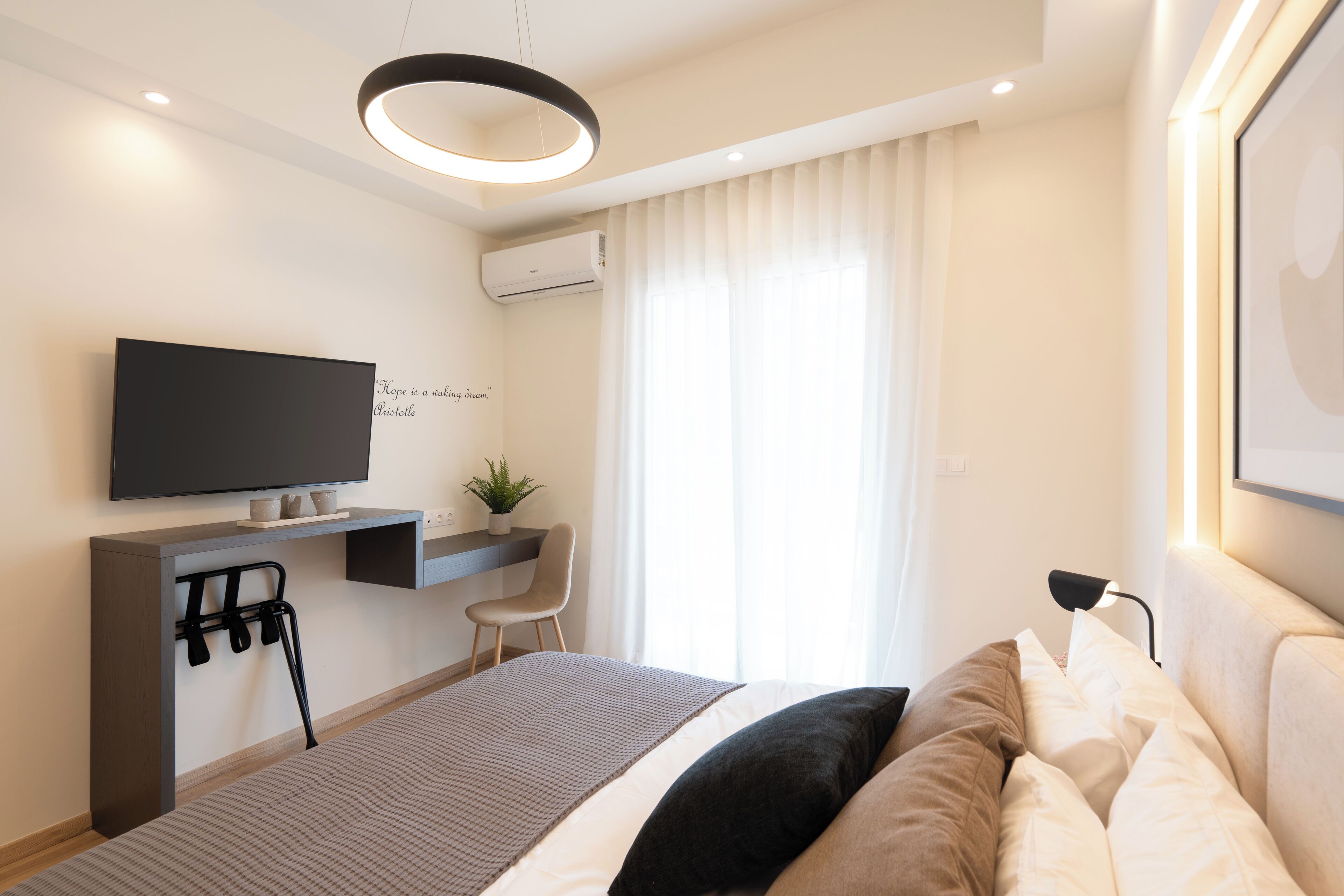 Ein modernes Hotelzimmer mit einem Bett, einem Schreibtisch mit Flachbildfernseher, einem Sessel und einem großen Fenster mit Vorhängen.
