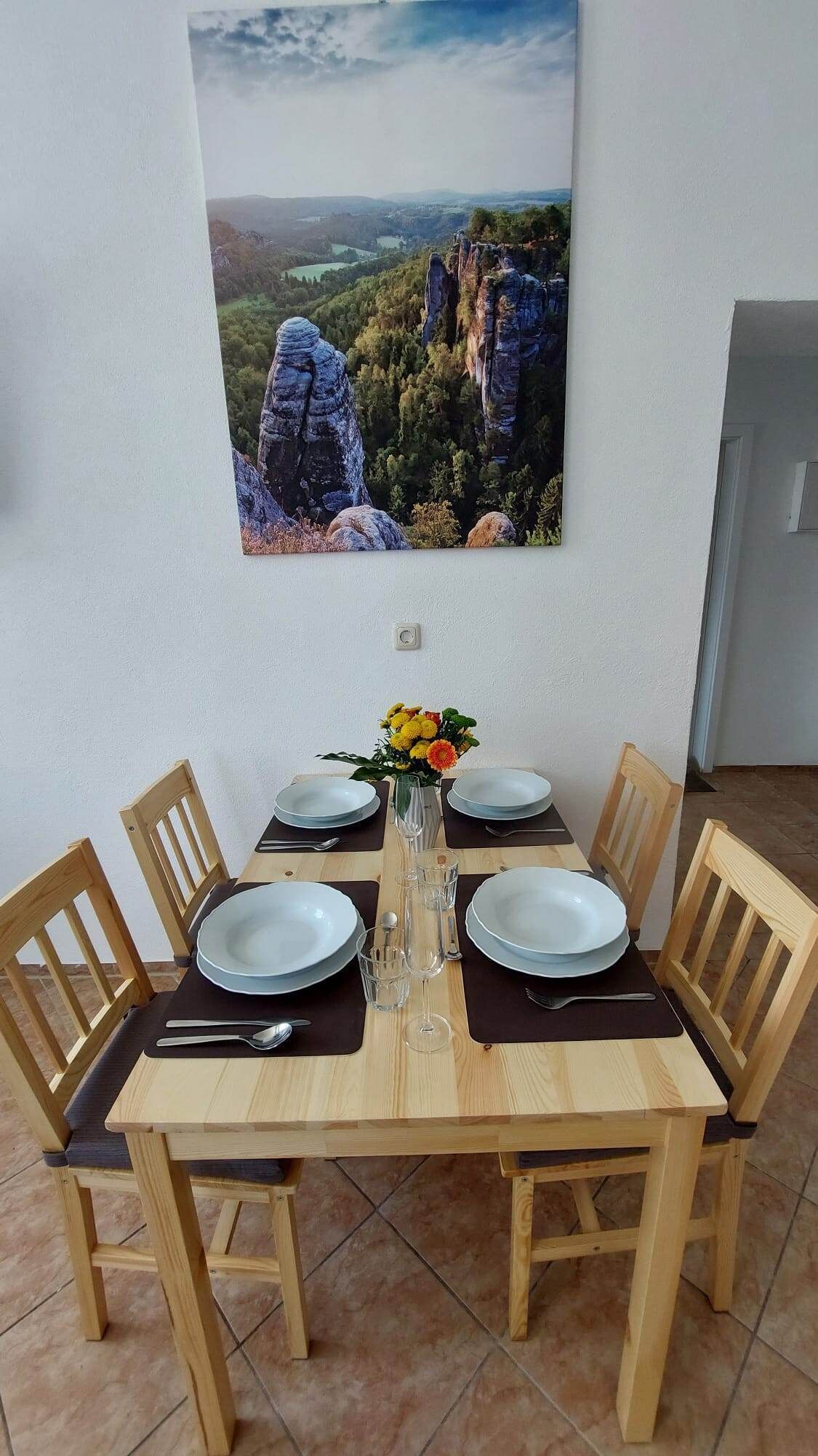 Ett matbord dukat med tallrikar, bestick och en vas med blommor, omgivet av trästolar, och ovanför hänger en vacker tavla som föreställer ett landskap.