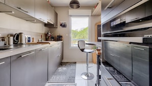 A modern kitchen with grey cabinets, a stainless steel kettle, a coffee machine, and a bar stool. - Escape at Les Sables d'Olonne Harbour – Sleeps 4 (Les Sables-d´Olonne)