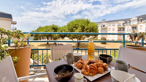 A balcony with a table set for breakfast, including croissants and a bottle of juice, overlooking a residential area with trees and buildings. - Escape at Les Sables d'Olonne Harbour – Sleeps 4 (Les Sables-d´Olonne)