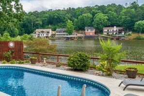 A swimming pool with a wooden deck, surrounded by a fence and a view of houses across a lake. - Serene Lakefront Escape with Pool & Hot Tub (Hot Springs)
