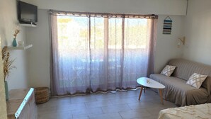 A room with a sofa, a small table, a television on a shelf, and a window with sheer curtains. - Studio for 4 with swimming pool and parking (Canet-en-Roussillon)