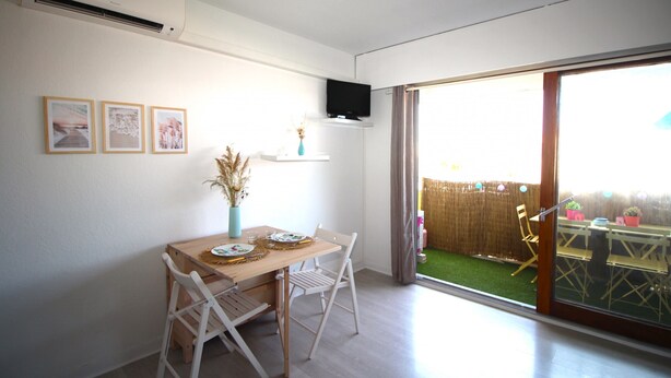 A dining area with a wooden table and white chairs, a TV mounted on the wall, and a sliding glass door leading to an outdoor patio with a wooden fence and potted plants. - Studio for 4 with swimming pool and parking (Canet-en-Roussillon)