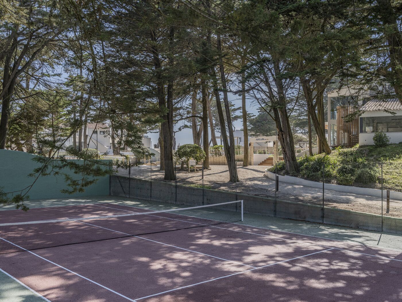 An outdoor tennis court surrounded by trees, with a building in the background.