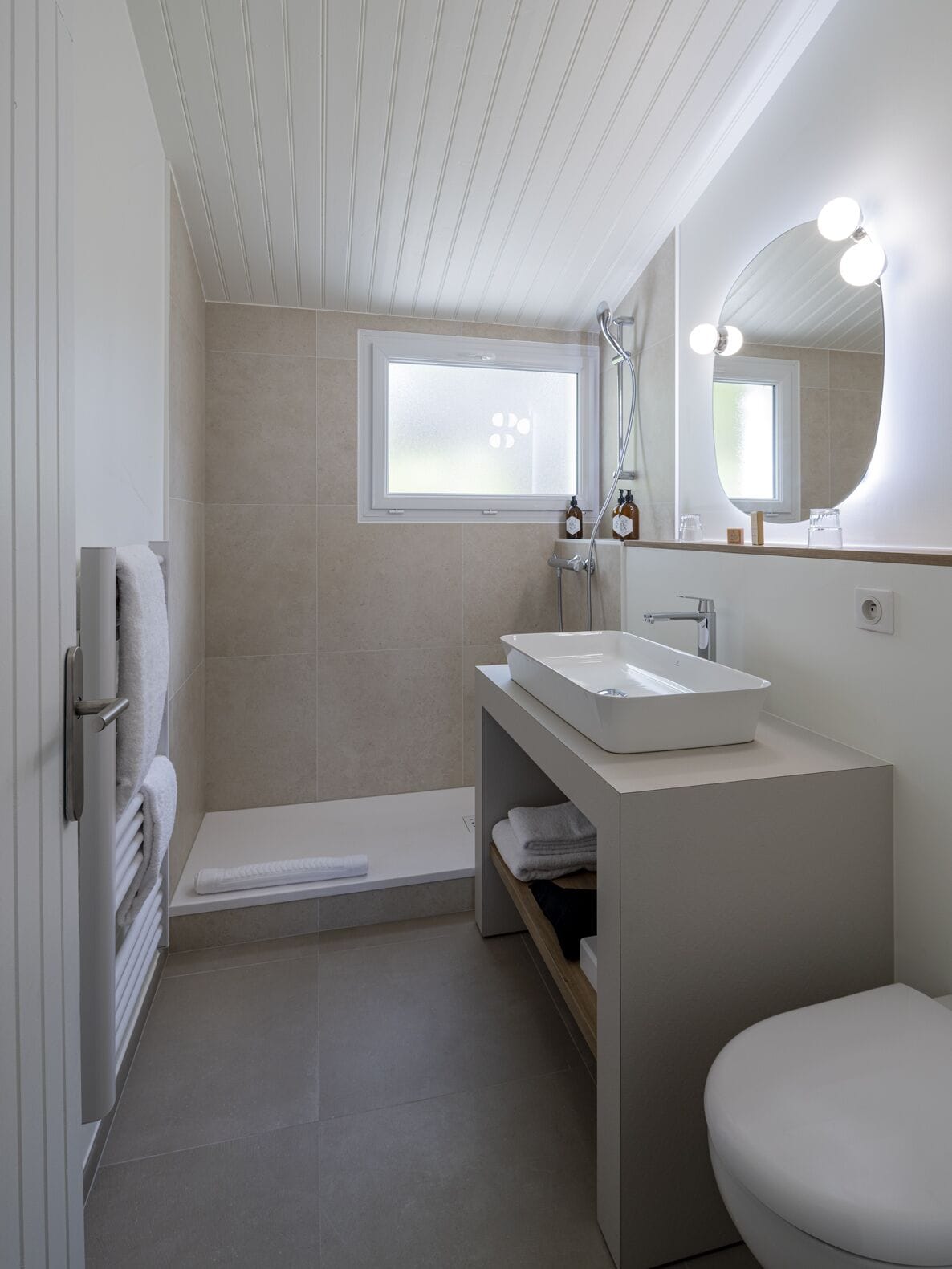 Ein modernes Badezimmer mit einem weißen Waschtisch, einem großen Spiegel mit Beleuchtung, einer Badewanne und einem Fenster.