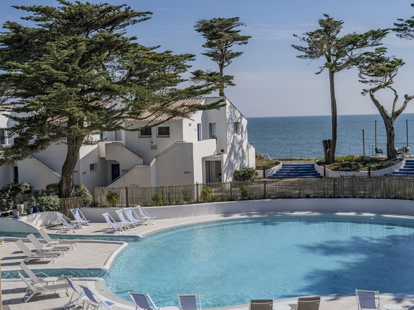 Ein Poolbereich mit Liegestühlen, freier Blick auf das Meer und ein Gebäude mit weißer Fassade.