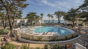 A circular pool with lounge chairs, surrounded by trees and a wooden deck, overlooking a beach and ocean. - Hôtel Punta Lara (La Gueriniere)