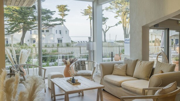 A modern interior with a sofa, chairs, a wooden table, and large windows offering a view of a white building and trees. - Hôtel Punta Lara (La Gueriniere)