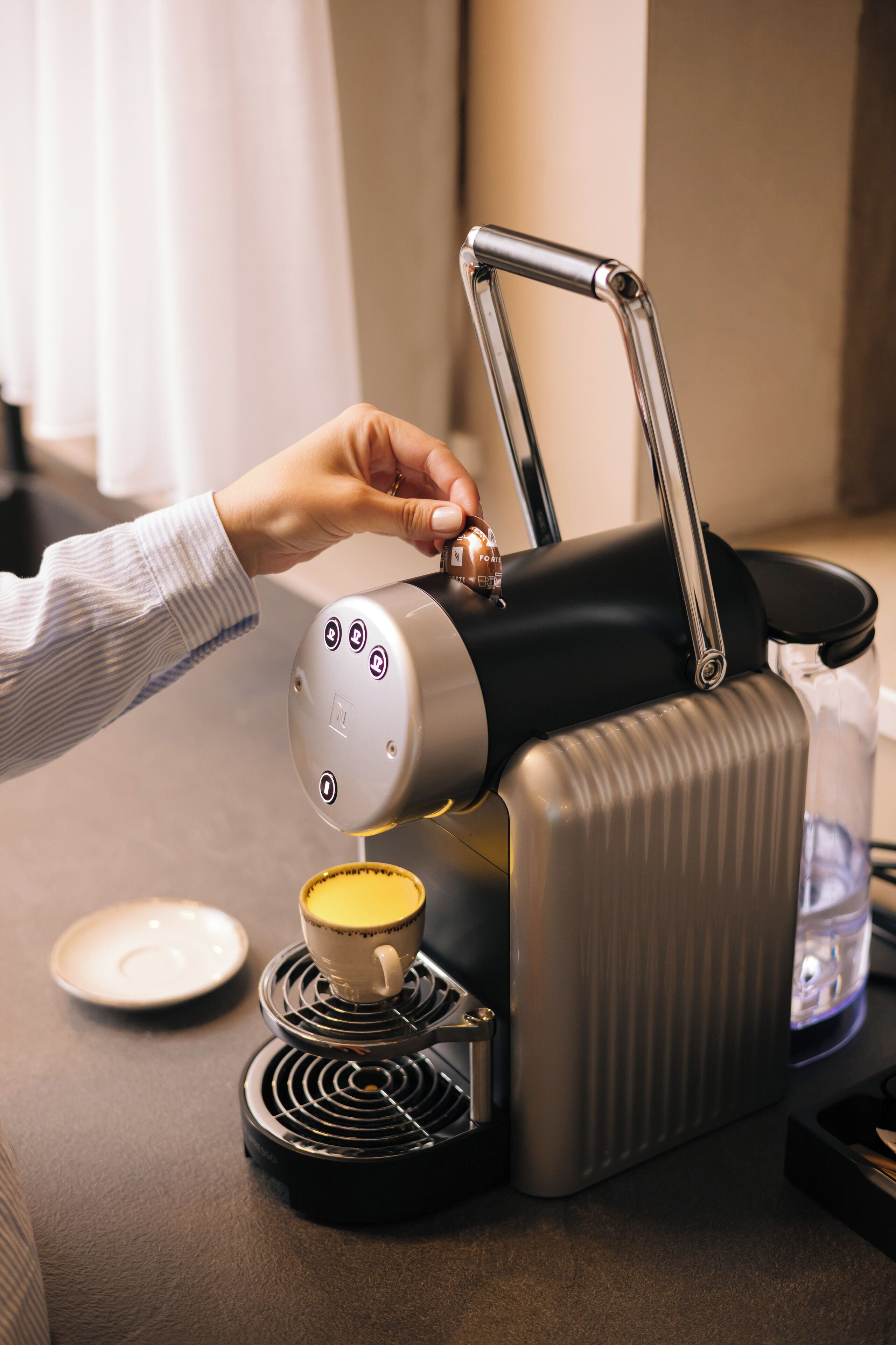 Eine Kaffeemaschine mit einer Tasse Kaffee auf einem Untersetzer, einem Teller und einer Wasserflasche auf dem Tisch.