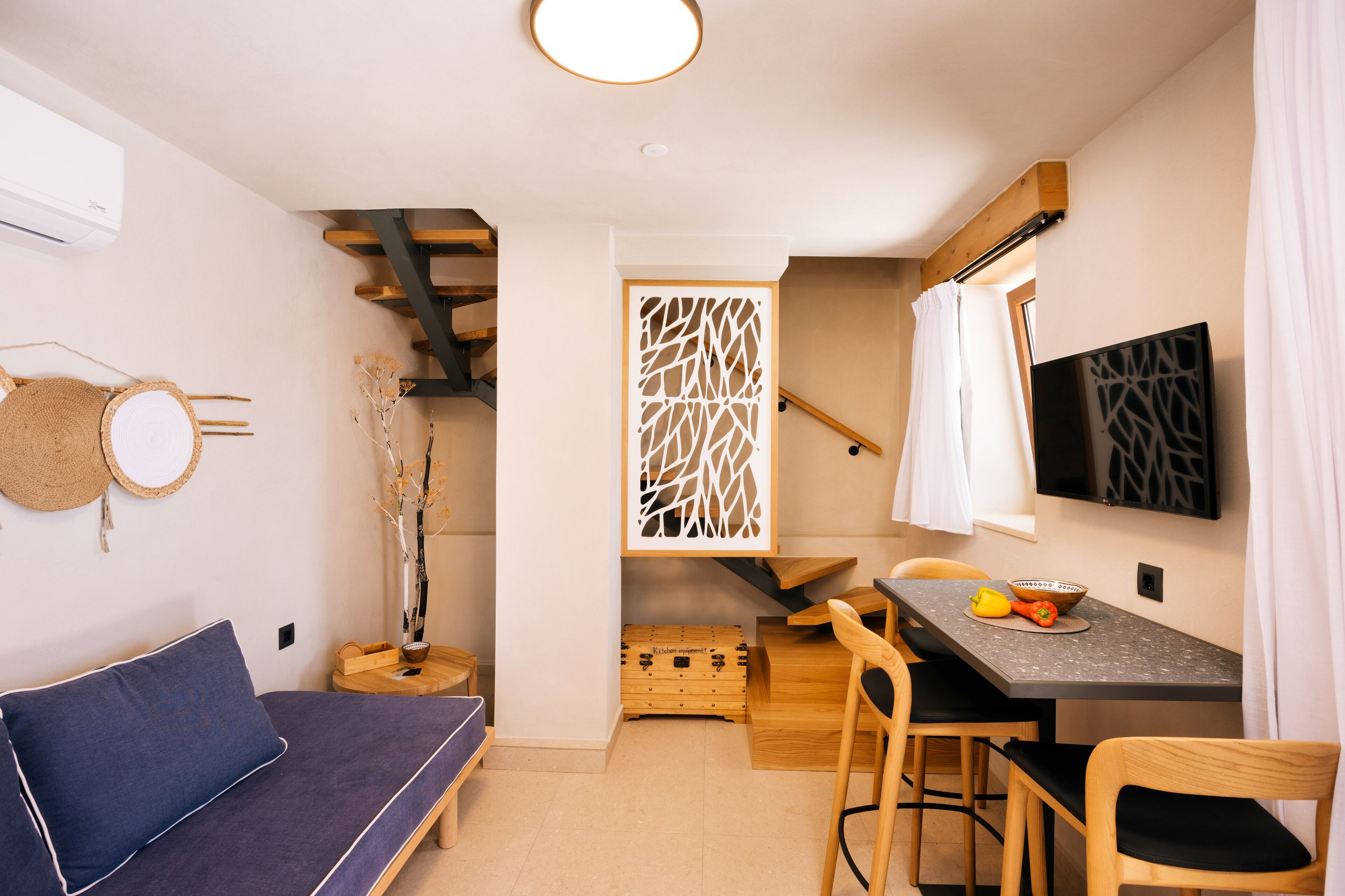 A compact living space with a sofa, a dining table with chairs, a TV mounted on the wall, and a staircase leading to an upper level.