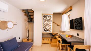 A compact living space with a sofa, a dining table with chairs, a TV mounted on the wall, and a staircase leading to an upper level.