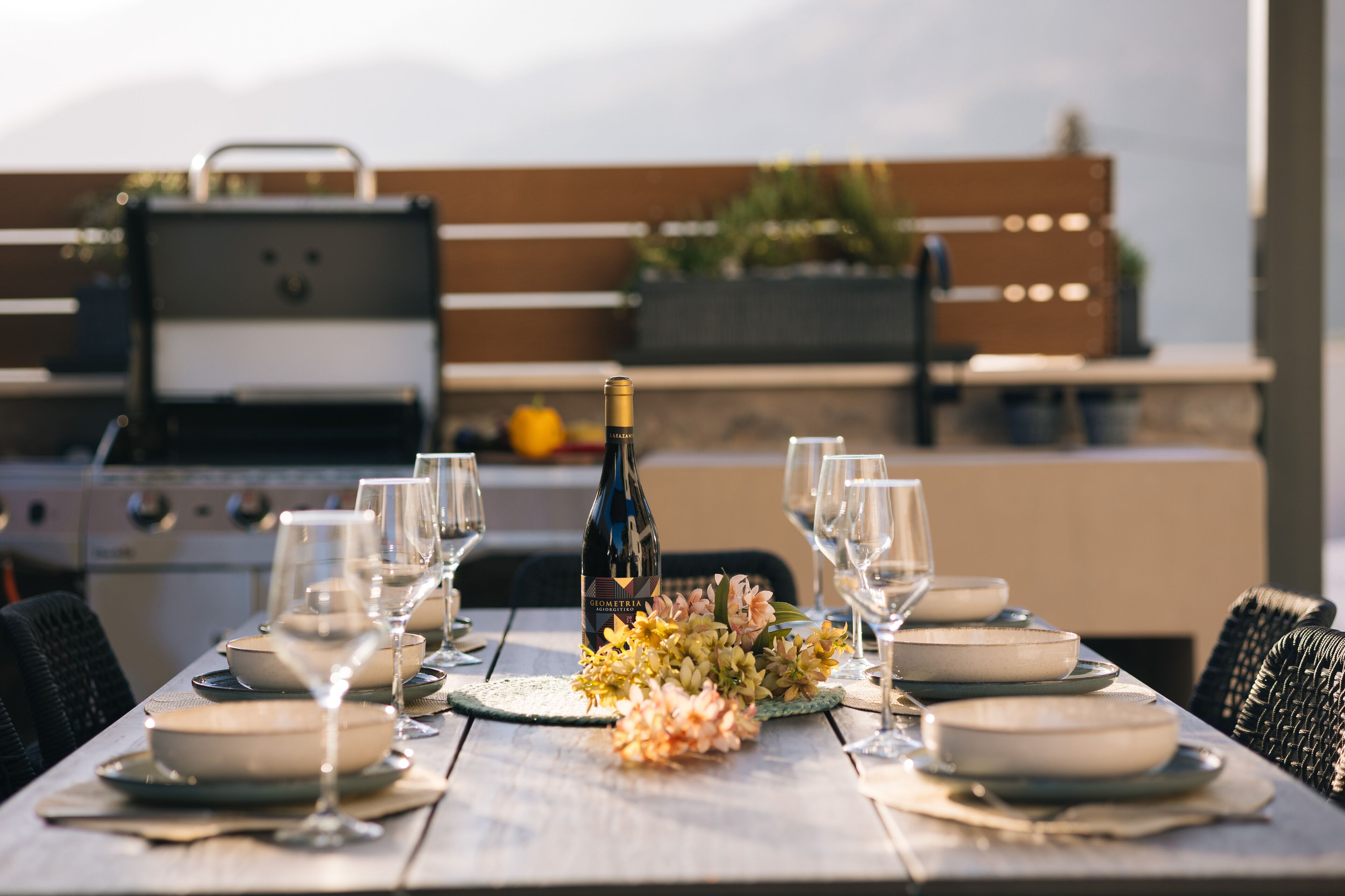 A dining table set for six with plates, bowls, and wine glasses, a bottle of wine, and a floral centerpiece.