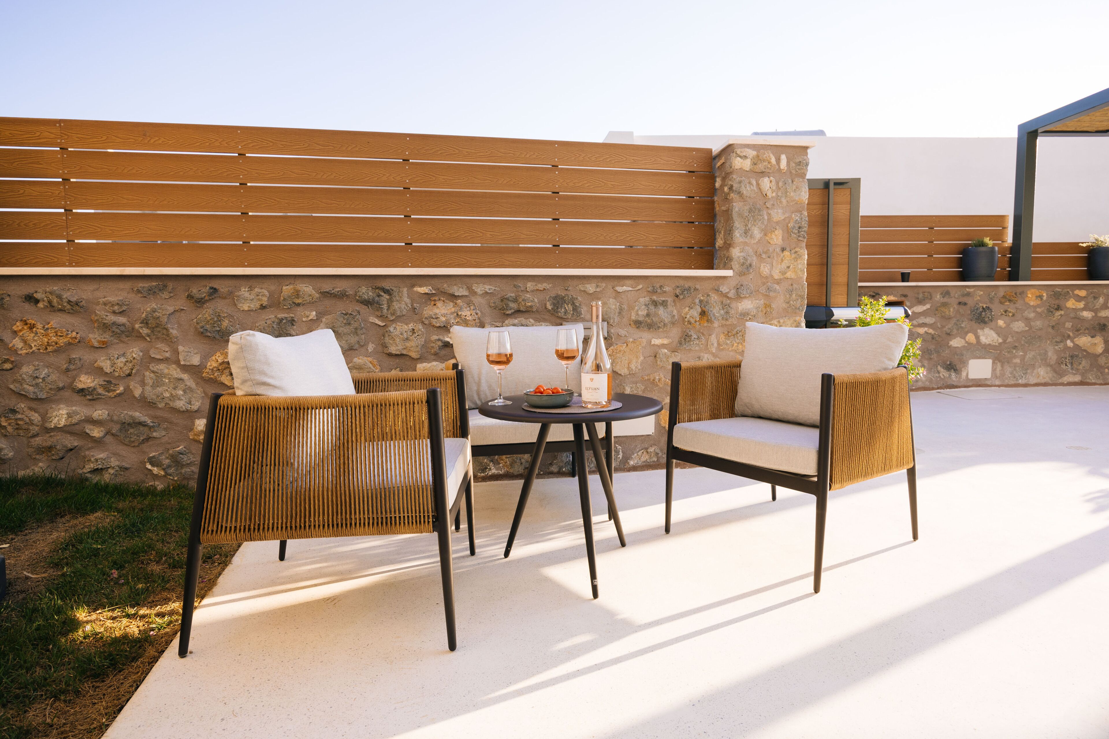 An outdoor seating area with wicker chairs and a round table, set against a stone wall and wooden slats.