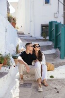 Un couple est assis sur un banc de pierre dans une ruelle étroite, bordée de plantes en pot et d’une clôture verte.