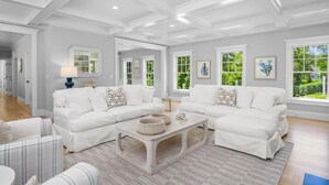 A spacious living room with white sofas, a coffee table, and large windows offering a view of greenery.