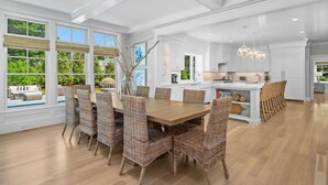 A spacious dining area with a large wooden table and wicker chairs, a kitchen with white cabinetry, and large windows offering a view of greenery.