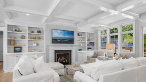 A spacious living room with white furniture, a fireplace, and large windows offering a view of the outdoors.