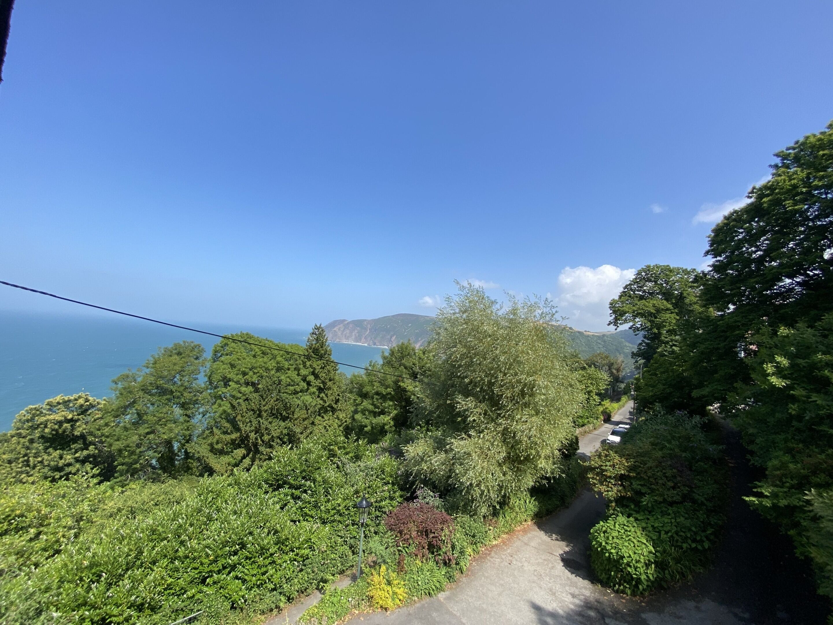 A pathway surrounded by dense greenery, leading to a scenic overlook with a view of the sea and distant hills.