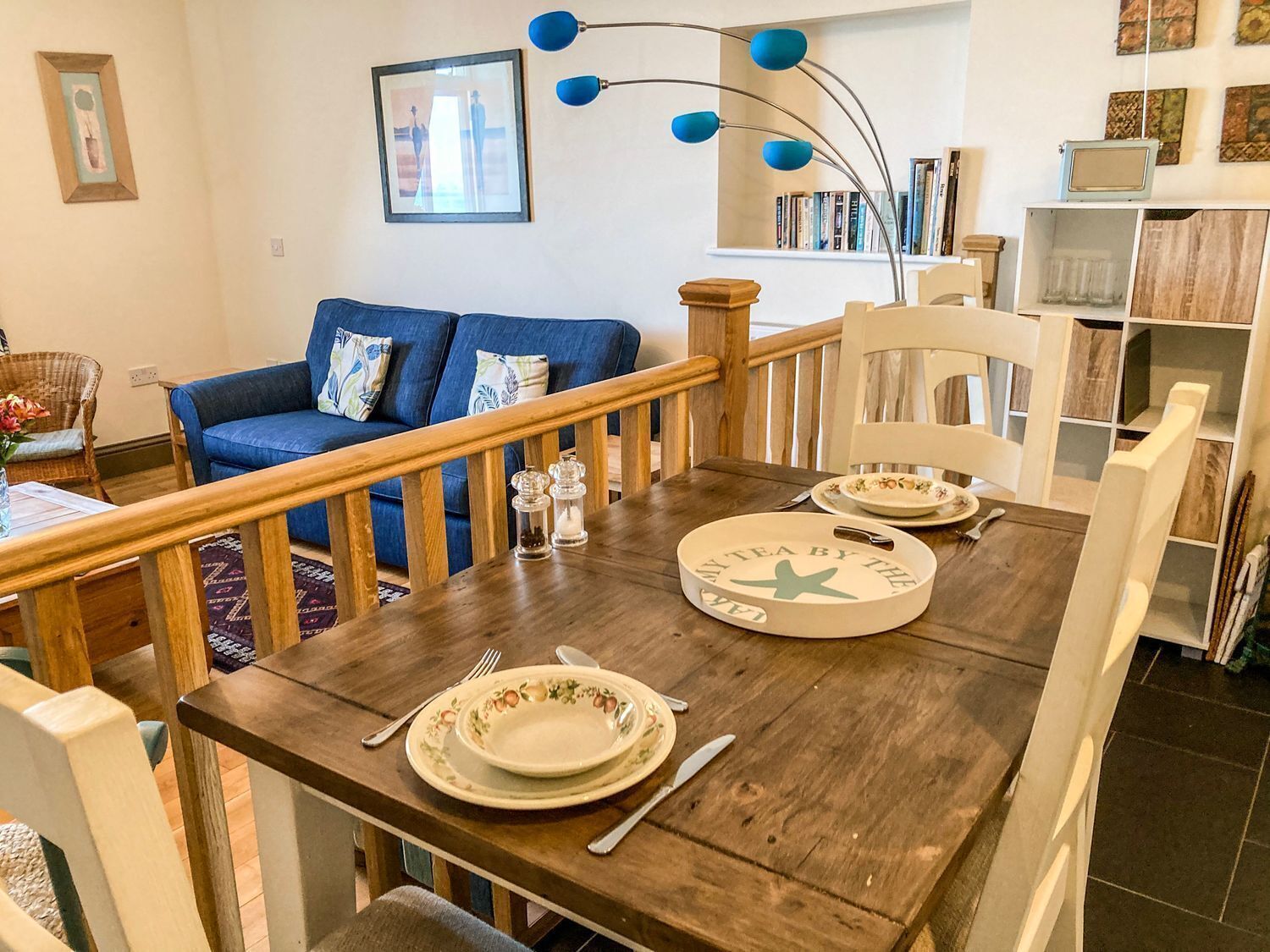 A dining area with a wooden table set for two, a blue sofa in the background, and a bookshelf with books and decorative items.