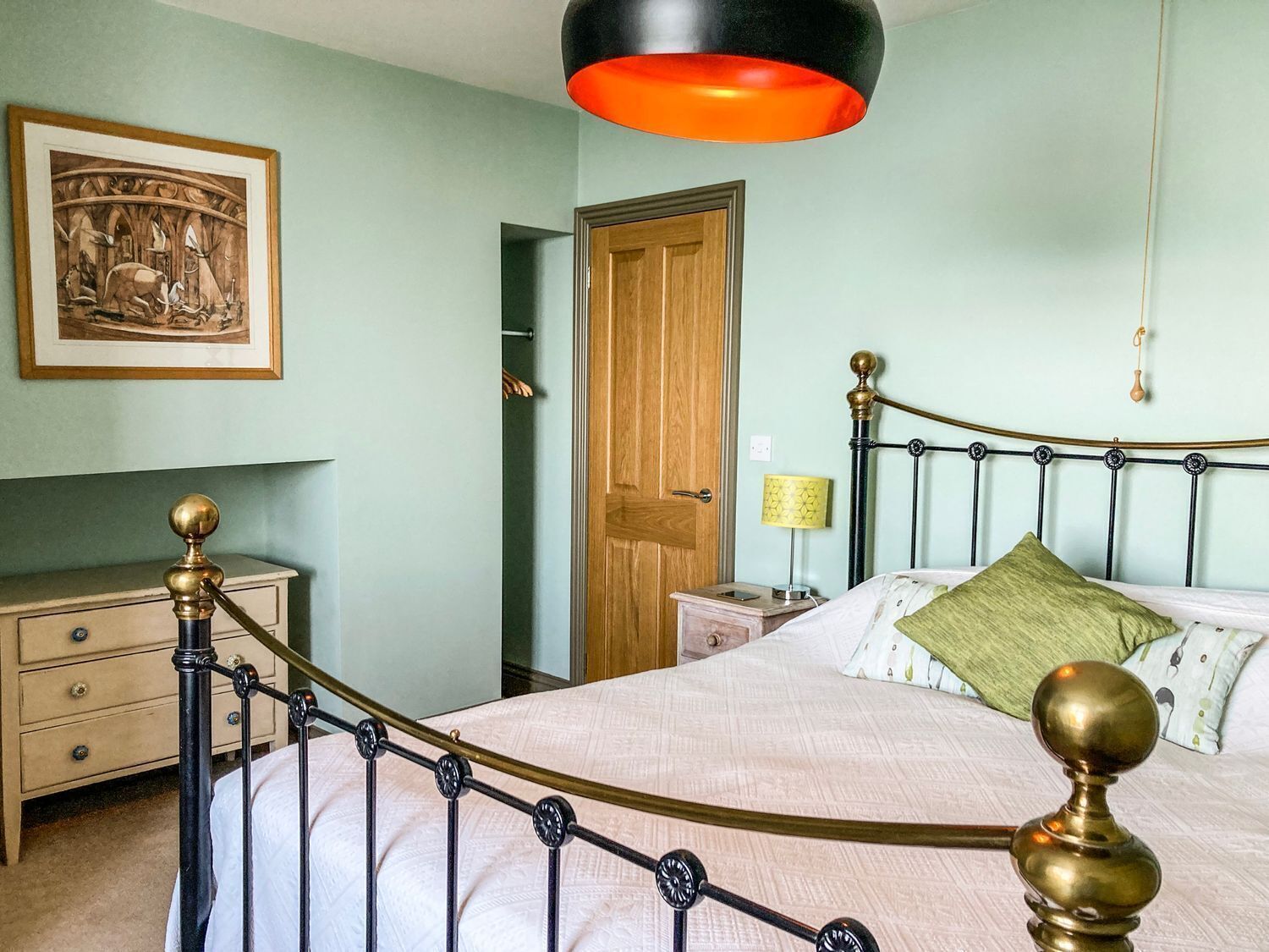 Een slaapkamer met een bed, nachtkastje, lamp, deur en een lijst met een afbeelding aan de muur.
