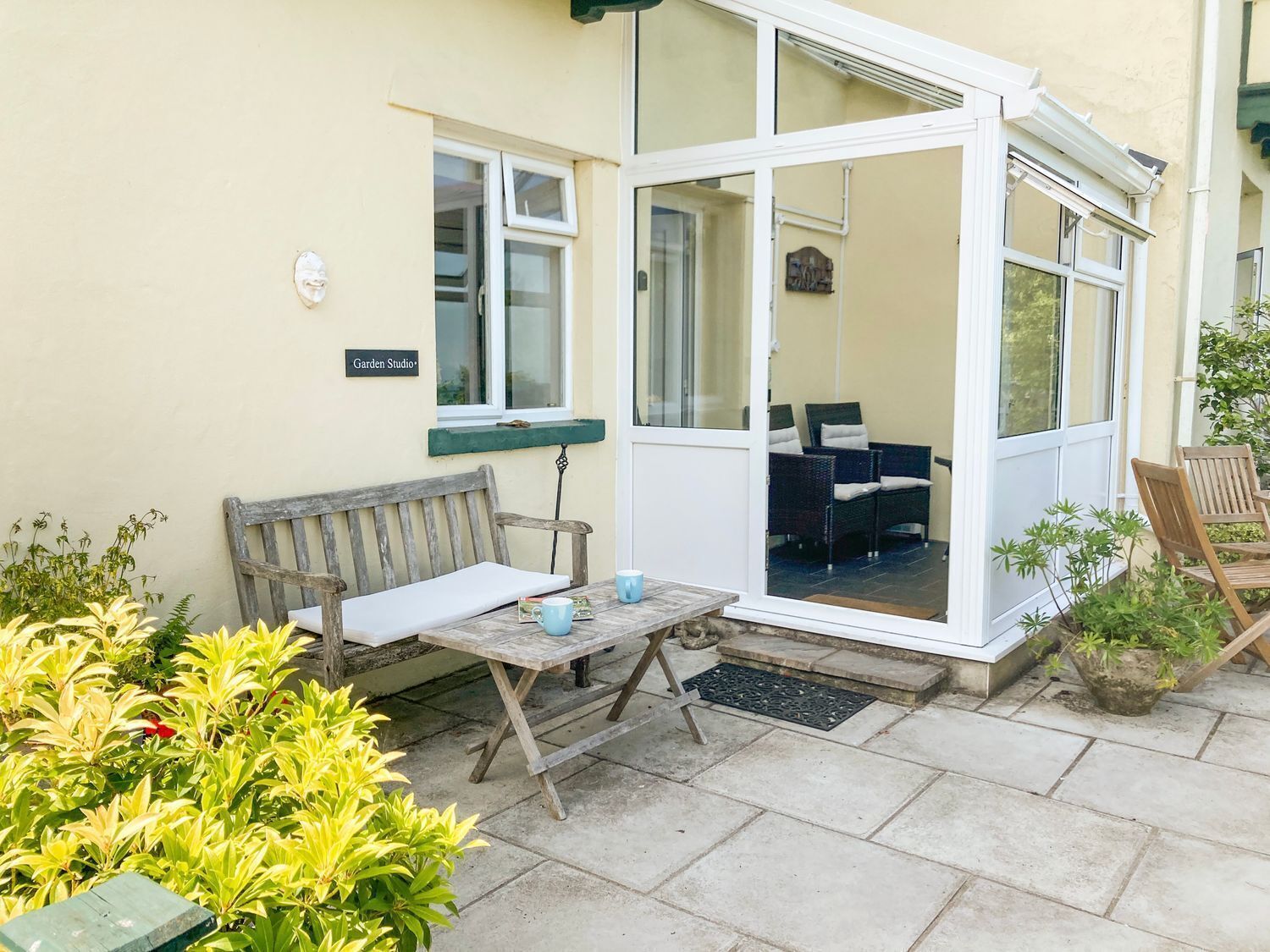 A patio area with a wooden bench, a table with two cups, and a glass door leading to a seating area with wicker furniture.