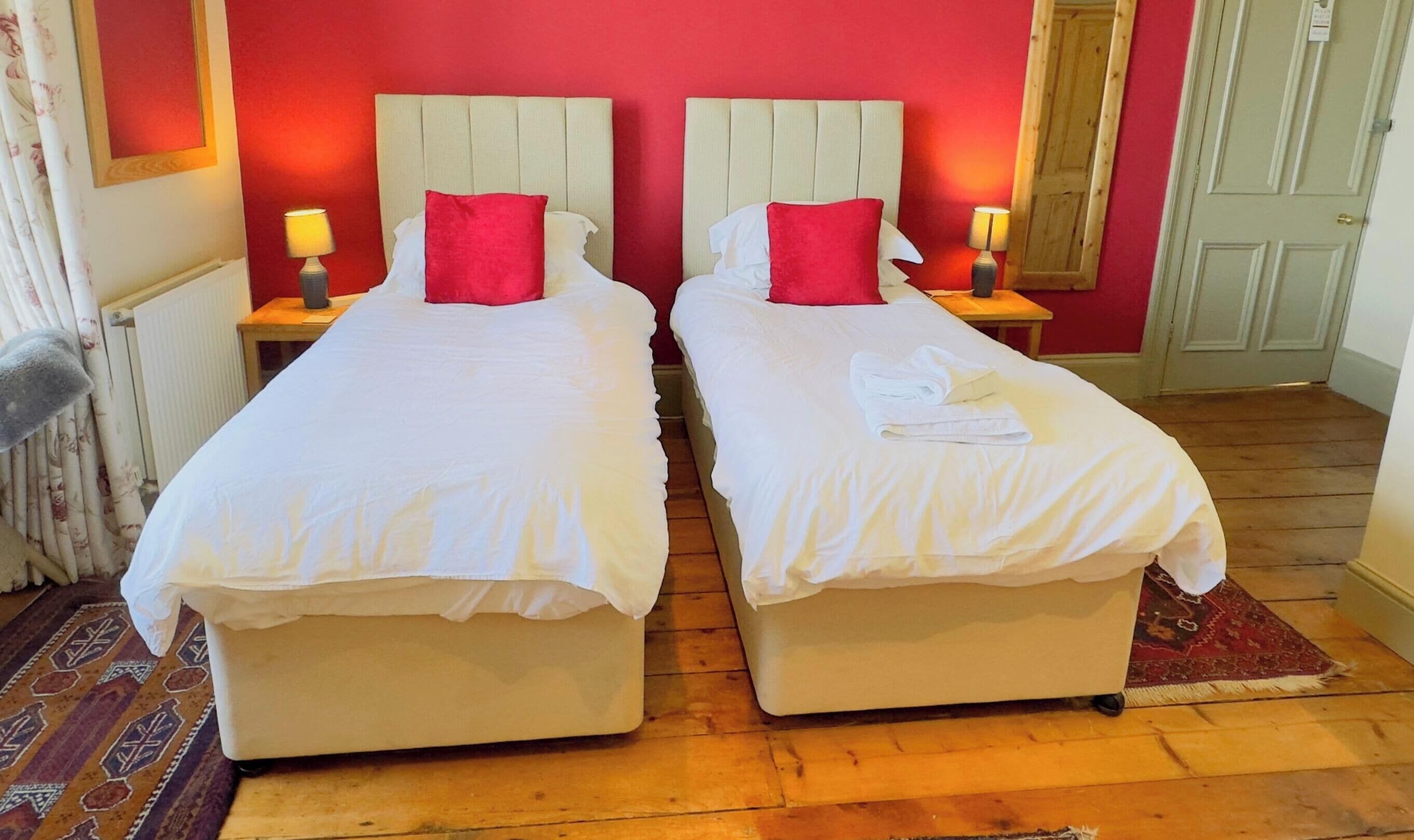 A room with two single beds, each with a red pillow and white bedding. There are two bedside tables with lamps, a radiator, and a patterned rug on the floor.